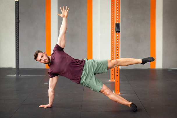Vue latérale de la forme masculine exécutant l'exercice de planche latérale d'étoile de calisthénique sur le sol dans la salle de gym moderne - Photo, image
