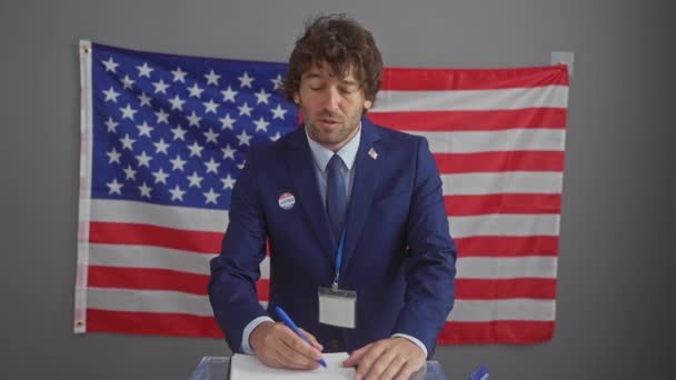 Ένας νεαρός άνδρας με μπλε κοστούμι ψηφίζει σε ένα εσωτερικό εκλογικό κέντρο της Αμερικής, με μια σημαία μας στο παρασκήνιο. - Πλάνα, βίντεο