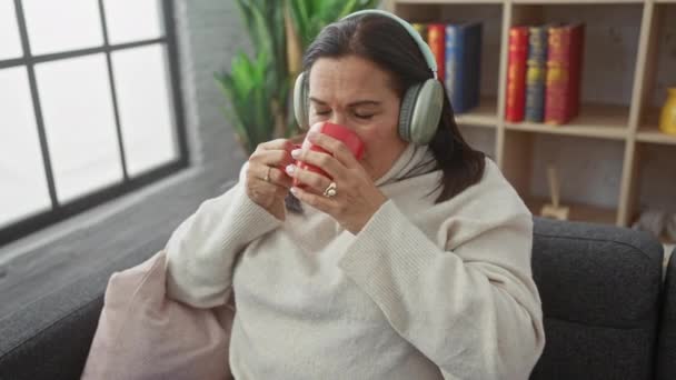 Een vrouw van middelbare leeftijd geniet van haar koffie thuis terwijl ze naar muziek luistert met een koptelefoon in een gezellige woonkamer.. - Video
