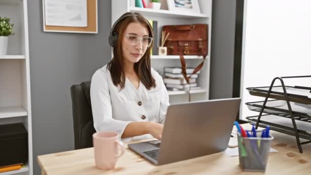 Εστιασμένη νεαρή γυναίκα φορώντας ακουστικά χρησιμοποιώντας φορητό υπολογιστή στο σύγχρονο γραφείο - Πλάνα, βίντεο