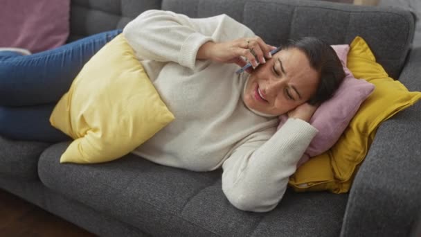 Een vrouw van middelbare leeftijd lacht terwijl ze telefoneert, liggend op een bank met kleurrijke kussens in een gezellige woonkamer. - Video