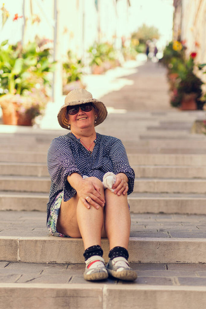 An old woman in a hat sits on the steps of a long staircase. A long street with steps is lined with potted plants, an old traveler smiles while sitting on the steps on a sunny summer day. Grandma is a tourist traveling around Europe. - Foto, Imagen