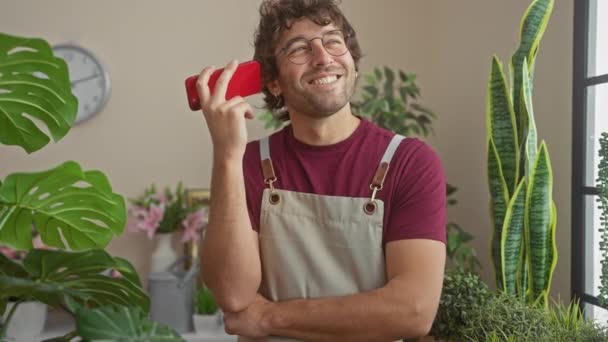 Homem feliz com barba vestindo avental sorri enquanto segurando o telefone na vibrante loja de plantas indoor. - Filmagem, Vídeo