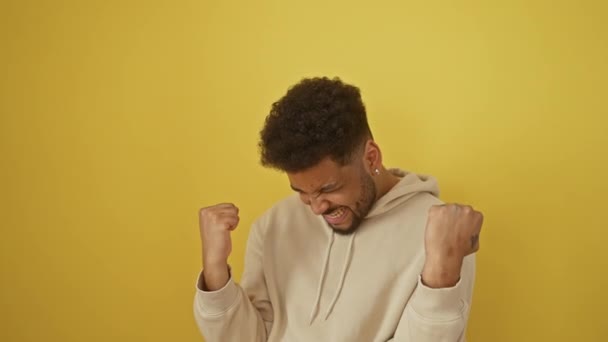 Victorious fun, cheerful african american man wearing hoodie screams in excited celebration of achievement. overjoyed winner expresses joy, confidence on isolated yellow background. - Footage, Video