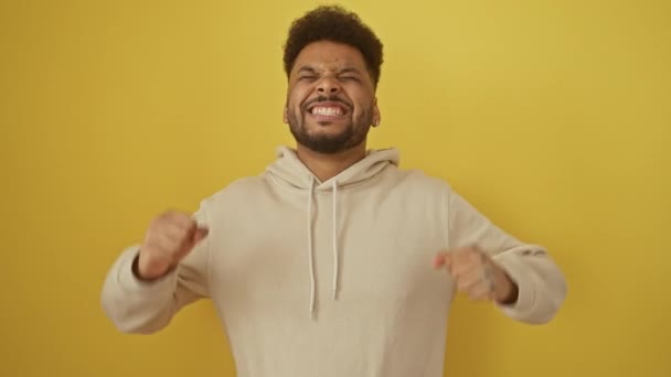 Opgewonden Afro-Amerikaanse man in een hoodie gek geworden van vreugde, het vieren van een gekke succes overwinning met armen wijd gegooid! winnaar vibes op een gele geïsoleerde achtergrond. - Video