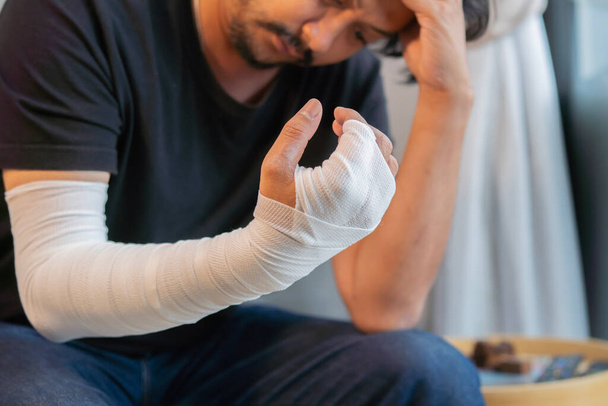 Homme asiatique adulte avec bandage de gaze enroulé autour de ses bras blessés assis à la maison avec préoccupation visage. Concentration sélective. - Photo, image