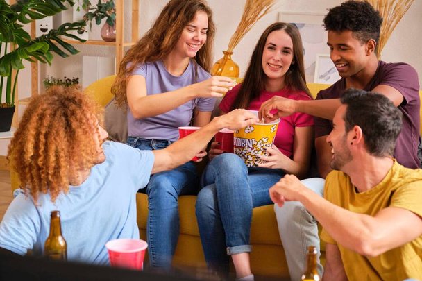 Πολυφυλετική ομάδα φίλων που τρώνε ποπ κορν ενώ βλέπουν ταινία σε κοινόχρηστο φοιτητικό διαμέρισμα. - Φωτογραφία, εικόνα
