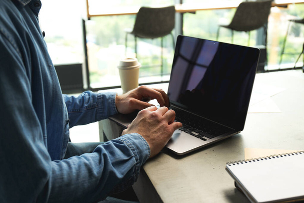 Чоловік працює ноутбуком, чашкою кави і блокнотами на столі в коворкінгу, крупним планом - Фото, зображення