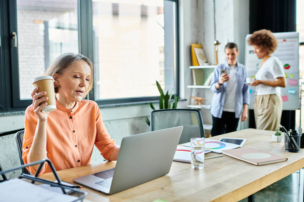 İşine dalmış bir kadın modern bir ofis alanında dizüstü bilgisayarın önünde çeşitli iş arkadaşlarıyla arka planda oturuyor.. - Fotoğraf, Görsel