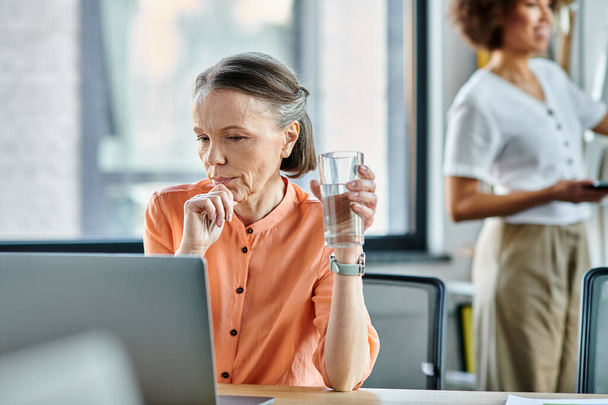 Toegewijde vrouw gericht op haar laptop, ondergedompeld in werk in een moderne kantooromgeving, met haar Afrikaanse Amerikaanse collega op de achtergrond. - Foto, afbeelding