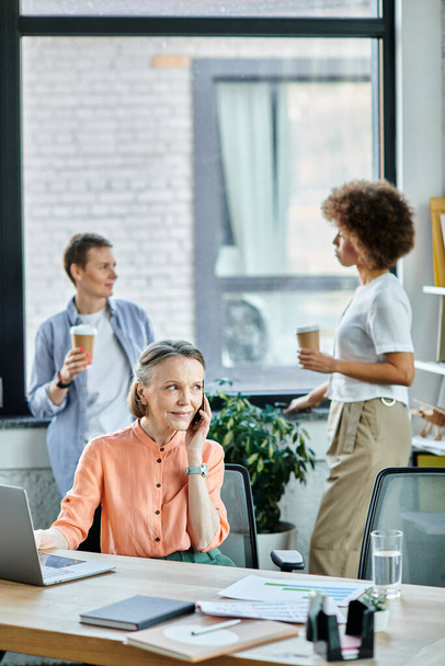 Devoted businesswoman talking on phone while using laptop, with her diverse colleagues on backdrop. - Photo, Image