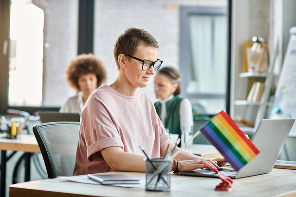 Appealing woman engrossed in work, with a laptop in front of her, with her diverse colleagues on backdrop, pride flag. - Photo, Image
