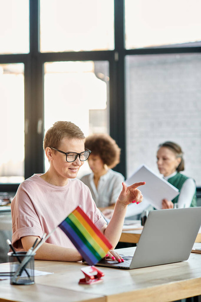Short haired woman engrossed in work, with a laptop in front of her, with her diverse colleagues on backdrop, pride flag. - Photo, Image