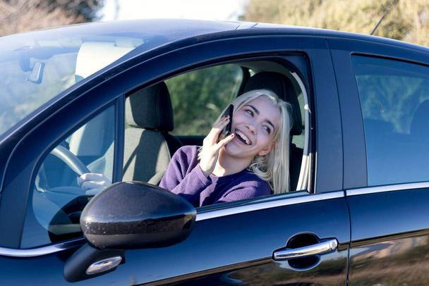 Egy mosolygó szőke nő lila pulóverben, aki a telefonján cseveg, miközben egy autó vezetőülésén ül. - Fotó, kép