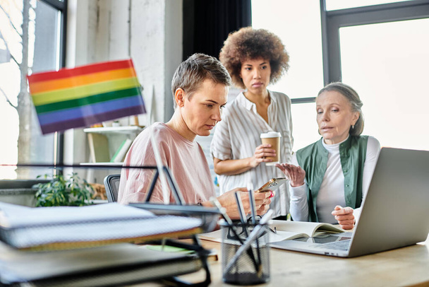 Mujeres de negocios atentas y diversas, incluyendo miembros de la comunidad LGBT, trabajando intensamente alrededor de una computadora portátil en una oficina. - Foto, Imagen