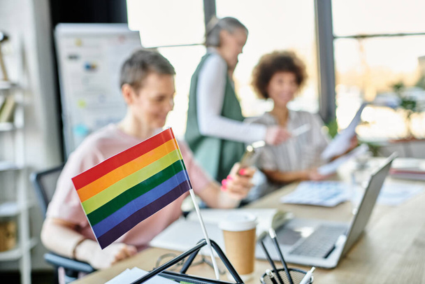 Enfoque en la bandera del arco iris que se muestra en la mesa de la oficina frente a las mujeres de negocios borrosas dverse. - Foto, Imagen