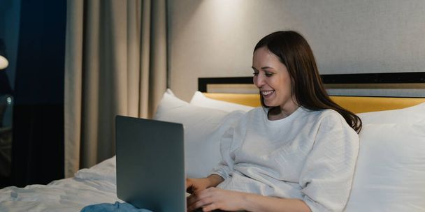 Donna felice utilizzando il computer portatile in camera da letto confortevole. - Foto, immagini