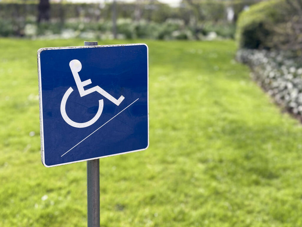 Un pequeño letrero azul para discapacitados en silla de ruedas se coloca sobre un fondo borroso del jardín, simbolizando la accesibilidad y la inclusividad en espacios al aire libre. - Foto, Imagen