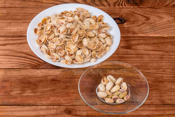 Partly peeled roasted salted pistachio nuts on the glass saucer against the empty shells on a big dish on a rustic table - Photo, Image