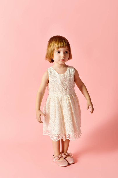 Маленька дівчинка в білій сукні витончено стоїть на фоні рожевого кольору. - Фото, зображення