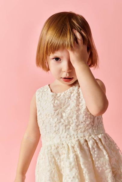 Urocza dziewczynka w białej sukience uderzające poza na miękkim różowym tle. - Zdjęcie, obraz
