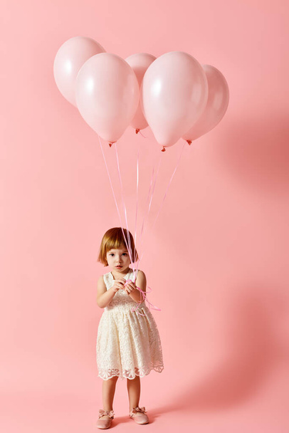 Αξιολάτρευτο κορίτσι σε λευκό φόρεμα κρατώντας ροζ μπαλόνια σε ροζ φόντο. - Φωτογραφία, εικόνα
