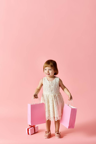 Beyaz elbiseli küçük bir kız pembe arka planda pembe alışveriş torbaları tutuyor.. - Fotoğraf, Görsel
