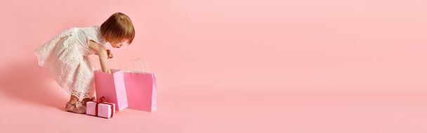 Adorable girl in white dress holding a vibrant pink shopping bag against a pink background. - Photo, Image