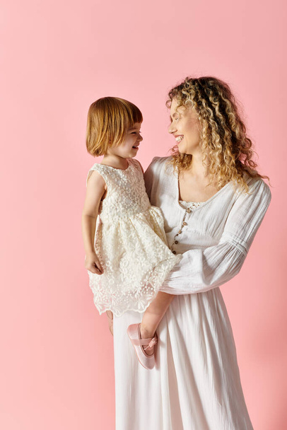 krullend haar moeder en schattig meisje in witte jurk staan voor een levendige roze achtergrond. - Foto, afbeelding