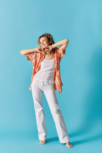 Elegante dame met krullend haar staat vol vertrouwen in witte broek en oranje shirt op levendige blauwe achtergrond. - Foto, afbeelding
