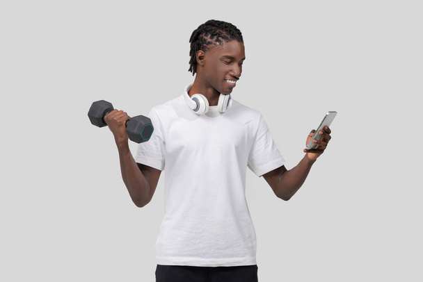 Přizpůsobit Afroameričan muž ležérně drží činku při pohledu na displej smartphone na bílém pozadí - Fotografie, Obrázek