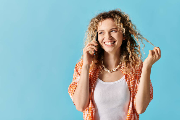 Νεαρή γυναίκα με σγουρά μαλλιά μιλάει στο τηλέφωνο σε μπλε φόντο. - Φωτογραφία, εικόνα