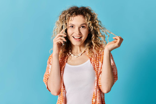 Una giovane donna con i capelli ricci che parla sul suo cellulare su uno sfondo blu vibrante. - Foto, immagini