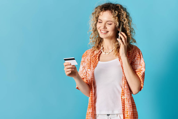 Młoda kobieta z kręconymi włosami trzyma kartę kredytową i rozmawia przez telefon, wszystko na tętniącym życiem tle. - Zdjęcie, obraz