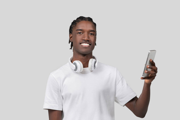 Šťastný mladý Afroameričan muž s úsměvem při používání svého smartphonu, nošení sluchátek na bílém pozadí - Fotografie, Obrázek