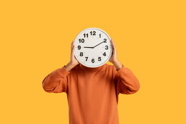 Ein Mann in orangefarbenem Pullover bedeckt sein Gesicht mit einer Wanduhr, die Begriffe wie Zeit und Fristen symbolisiert. - Foto, Bild