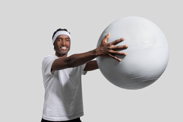 Urheileva nuori afroamerikkalainen mies aktiivisissa vaatteissa osoittaa rintakehää, jolla on suuri harjoituspallo tavallista taustaa vasten. - Valokuva, kuva