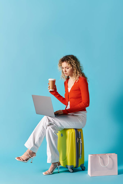 A woman with curly hair sits on a suitcase, sipping coffee and working on a laptop. - Photo, Image