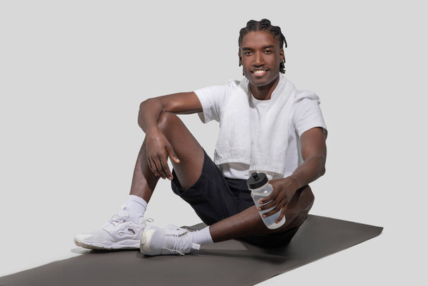 Χαμογελώντας και καθισμένος σε ένα στρώμα γιόγκα, ένας ενεργός Αφροαμερικάνος απολαμβάνει ένα διάλειμμα κρατώντας ένα διάφανο μπουκάλι νερό σε ένα στούντιο - Φωτογραφία, εικόνα