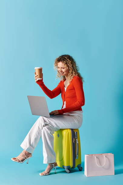 Curly-haired woman sits on suitcase with coffee and laptop. - Photo, Image