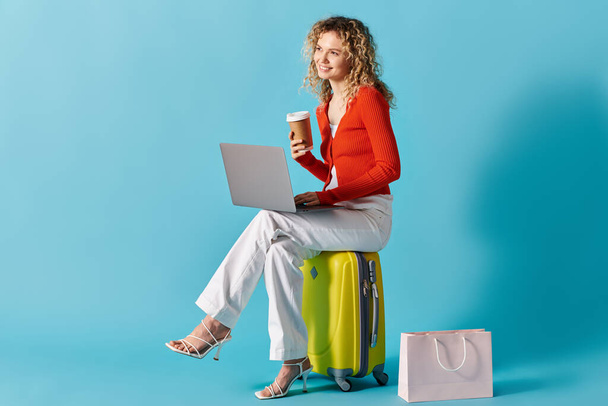 Kędzierzawa kobieta siedzi na walizce, pisze na laptopie i pije kawę.. - Zdjęcie, obraz