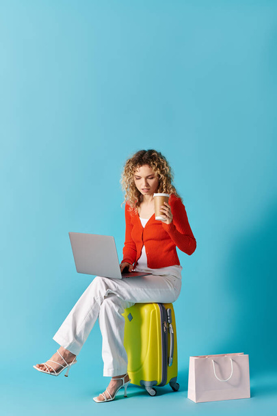 Curly-haired woman sits on suitcase, sipping coffee, using laptop. - Photo, Image