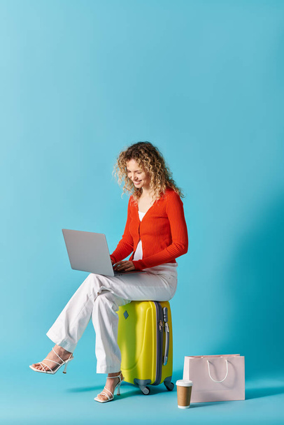 Kiharatukkainen nainen istuu matkalaukussa, kannettavan tietokoneen avulla. - Valokuva, kuva