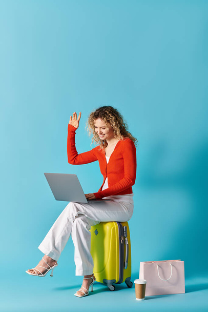 Kıvırcık saçlı kadın dizüstü bilgisayara dalmış bir şekilde bavulun üzerine oturur.. - Fotoğraf, Görsel