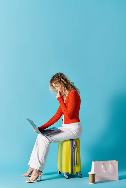 Eine Frau mit lockigem Haar sitzt auf einem Koffer und arbeitet an einem Laptop. - Foto, Bild