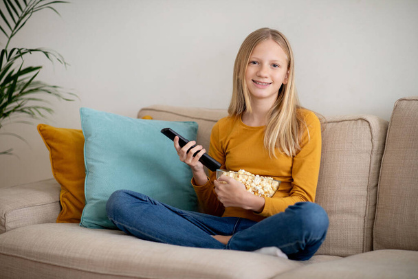 Χαρούμενη έφηβη που κρατάει ένα τηλεχειριστήριο και ένα μπολ ποπ κορν, έτοιμη να απολαύσει ένα χαλαρωτικό βράδυ στον καναπέ, βλέποντας τηλεόραση στο σαλόνι - Φωτογραφία, εικόνα