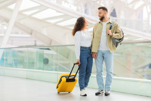 Happy young couple engaging in conversation while walking with a yellow suitcase at an airport, cheerful spouses travelling together, copy space - Photo, Image
