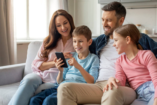 Sonriente familia sentada en el sofá en el acogedor interior de la casa como un niño les muestra algo en un teléfono inteligente - Foto, Imagen
