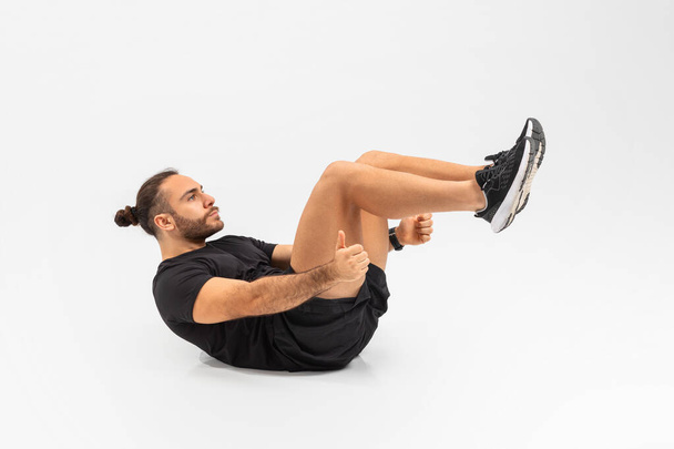 Energetischer Mann in schwarzer Sportkleidung, der vor schlichtem weißen Hintergrund eine schwebende Crunch-Übung durchführt - Foto, Bild