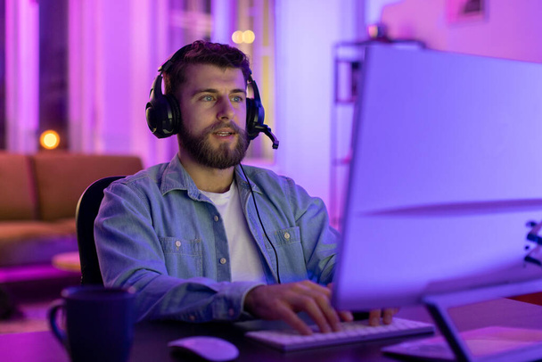 In una stanza con luci al neon, un uomo concentrato usa un auricolare mentre usa un computer con un'espressione seria. - Foto, immagini